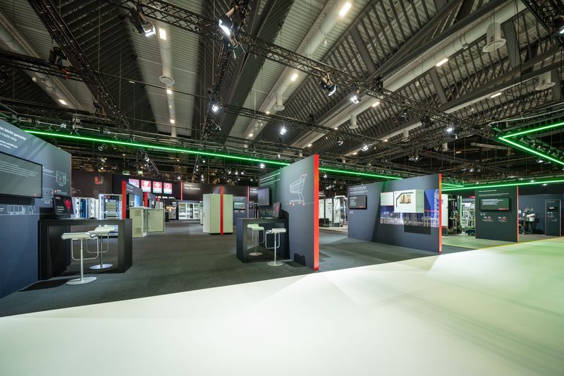 Messestand vor der Öffnung: Themeninseln, dunkle Wandscheiben mit roten Lichtkanten, weiße Lichtwege, grünes LED-Rigg