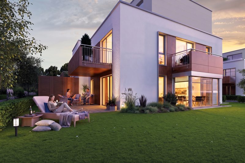 Visualisierung, 2026 mit KI veredelt und belebt: kubisches Wohnhaus mit Garten in der Abenddämmerung, eine Frau liest auf der Liege, zwei Personen auf der Terrasse